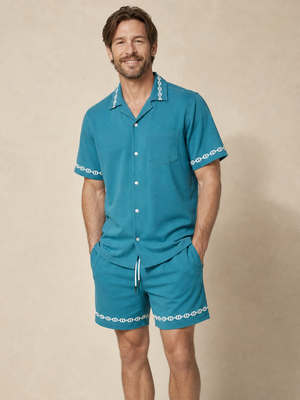 A man modeling a teal two-piece pajama set with white decorative trim on the collar, sleeve cuffs, and shorts, standing against a neutral beige background.