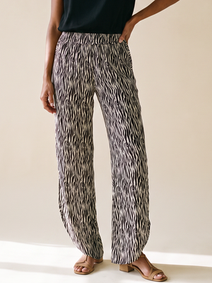A woman modeling a black short-sleeve top and high-waisted, wide-leg trousers with a black and white abstract pattern, standing against a warm beige studio background.
