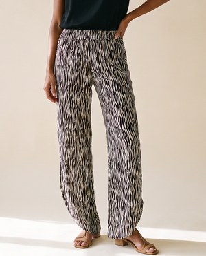 A woman modeling a black short-sleeve top and high-waisted, wide-leg trousers with a black and white abstract pattern, standing against a warm beige studio background.