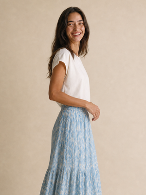 A woman modeling a light blue and white patterned tiered maxi skirt with a white short-sleeve top, standing against a neutral beige studio backdrop.