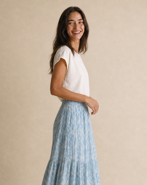 A woman modeling a light blue and white patterned tiered maxi skirt with a white short-sleeve top, standing against a neutral beige studio backdrop.