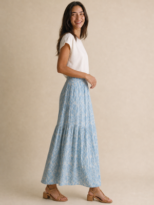 A woman modeling a light blue and white patterned tiered maxi skirt with a white short-sleeve top, standing against a neutral beige studio backdrop.
