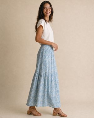 A woman modeling a light blue and white patterned tiered maxi skirt with a white short-sleeve top, standing against a neutral beige studio backdrop.
