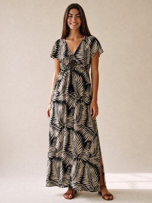 A woman modeling a black and beige palm leaf print maxi dress with short flutter sleeves, a V-neckline, and a tie detail at the waist, standing against a neutral beige studio backdrop.