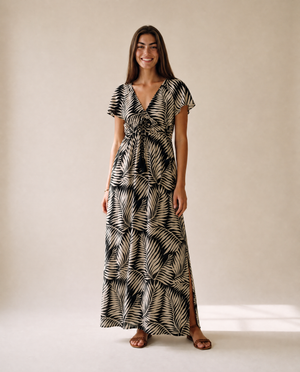 A woman modeling a black and beige palm leaf print maxi dress with short flutter sleeves, a V-neckline, and a tie detail at the waist, standing against a neutral beige studio backdrop.