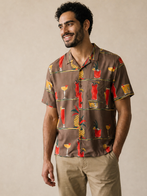 A man wearing a brown short-sleeve button-up shirt with a colorful cocktail and pineapple print, paired with beige pants and white sneakers, standing against a neutral beige wall.
