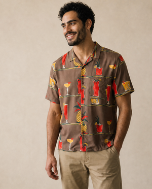 A man wearing a brown short-sleeve button-up shirt with a colorful cocktail and pineapple print, paired with beige pants and white sneakers, standing against a neutral beige wall.