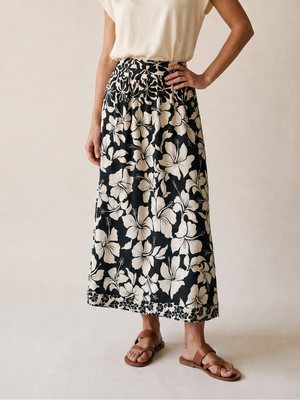 A woman modeling a cream short-sleeve top and a black midi skirt with a large cream floral print, standing against a plain beige studio background.