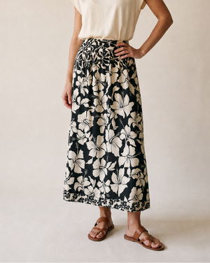 A woman modeling a cream short-sleeve top and a black midi skirt with a large cream floral print, standing against a plain beige studio background.