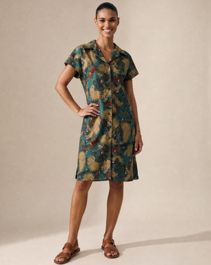 A woman modeling a short-sleeve button-front shirt dress with an abstract watercolor-style print in teal, olive green, mustard, and brown tones, paired with brown strappy sandals against a warm beige studio backdrop.