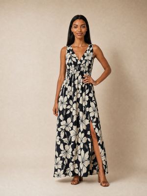 A woman modeling a sleeveless maxi dress with a black and off-white floral print, featuring a V-neckline, waist tie, and a high side slit, set against a plain beige studio backdrop.