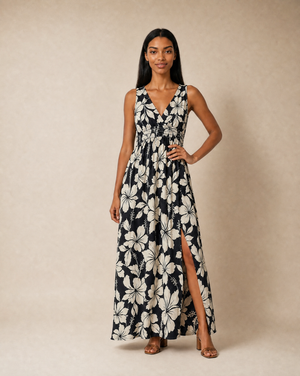 A woman modeling a sleeveless maxi dress with a black and off-white floral print, featuring a V-neckline, waist tie, and a high side slit, set against a plain beige studio backdrop.