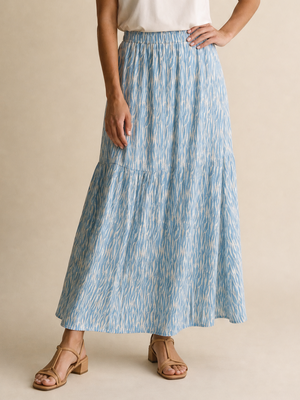 A woman modeling a white short-sleeve crewneck t-shirt and a high-waisted, tiered light blue maxi skirt with a subtle vertical abstract pattern, paired with tan strappy sandals, against a solid beige studio backdrop.