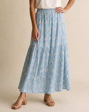 A woman modeling a white short-sleeve crewneck t-shirt and a high-waisted, tiered light blue maxi skirt with a subtle vertical abstract pattern, paired with tan strappy sandals, against a solid beige studio backdrop.
