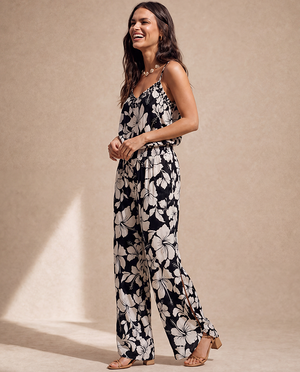 A woman modeling a sleeveless black jumpsuit with a large white floral print, standing against a textured beige studio backdrop.