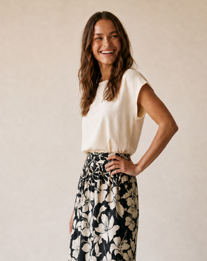 A woman modeling a cream short-sleeve top and a black maxi skirt with a large cream floral print, standing against a warm beige studio backdrop.
