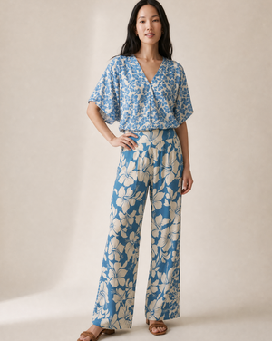 A woman modeling a matching blue floral top and wide-leg pants set against a warm beige studio backdrop.