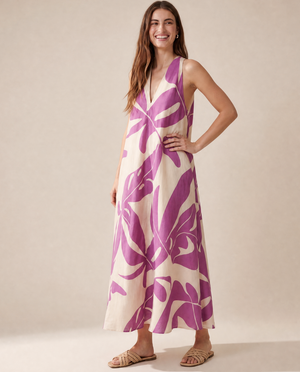 A woman modeling a sleeveless maxi dress with a bold purple tropical leaf pattern on a cream background, paired with tan flat sandals, against a solid beige studio backdrop.