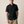 A man wearing a black short-sleeve button-up shirt and khaki shorts, standing against a plain beige studio background.