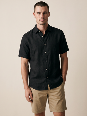 A man wearing a black short-sleeve button-up shirt and khaki shorts, standing against a plain beige studio background.