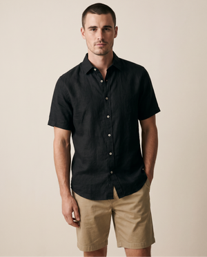 A man wearing a black short-sleeve button-up shirt and khaki shorts, standing against a plain beige studio background.