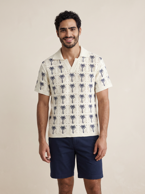A man wearing a short-sleeve camp collar shirt with a cream base and navy blue palm tree pattern, paired with navy blue shorts, standing against a solid beige background.