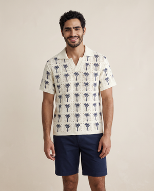 A man wearing a short-sleeve camp collar shirt with a cream base and navy blue palm tree pattern, paired with navy blue shorts, standing against a solid beige background.