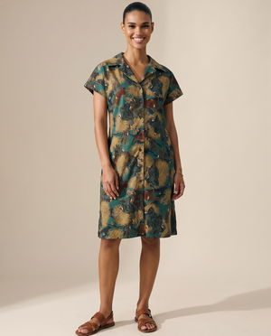 A woman modeling a short-sleeve button-front shirt dress with an abstract watercolor-style print in teal, olive green, tan, and brown tones, paired with brown leather sandals, against a plain beige studio background.