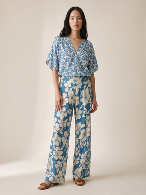 A woman modeling a matching two-piece set featuring a blue and beige floral print, consisting of a short-sleeve V-neck top and wide-leg trousers, set against a plain beige studio background.