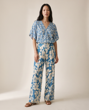 A woman modeling a matching two-piece set featuring a blue and beige floral print, consisting of a short-sleeve V-neck top and wide-leg trousers, set against a plain beige studio background.
