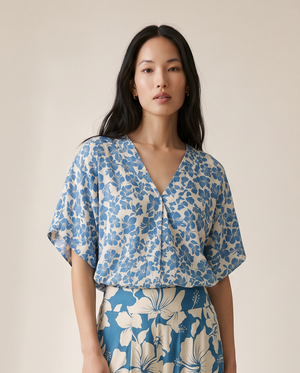 A woman modeling a matching two-piece set featuring a blue and beige floral print, consisting of a short-sleeve V-neck top and wide-leg trousers, set against a plain beige studio background.