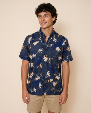 A man wearing a navy blue short-sleeve button-up shirt with a white and beige floral pattern, paired with khaki shorts, standing against a solid beige background.