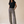 A woman stands in a studio wearing a black short-sleeve top and high-waisted, wide-leg trousers with an abstract black, white, and gray pattern. She is wearing tan heeled sandals against a warm beige background.