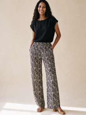 A woman stands in a studio wearing a black short-sleeve top and high-waisted, wide-leg trousers with an abstract black, white, and gray pattern. She is wearing tan heeled sandals against a warm beige background.