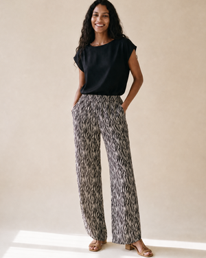A woman stands in a studio wearing a black short-sleeve top and high-waisted, wide-leg trousers with an abstract black, white, and gray pattern. She is wearing tan heeled sandals against a warm beige background.